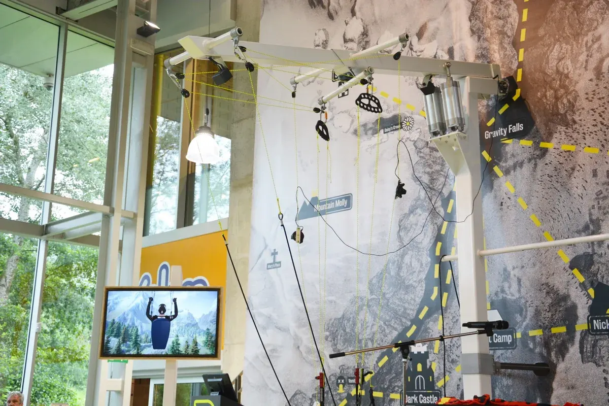 Technopolis paragliding installation with suspended harness equipment, a climbing-wall backdrop and a display screen.